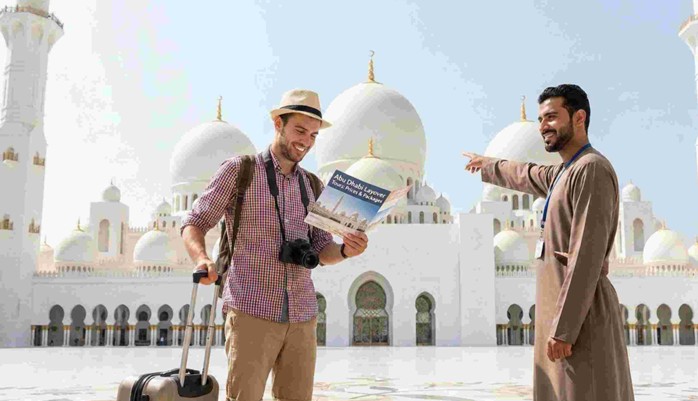 A tourist with a suitcase holds a brochure for 'Abu Dhabi Layover Tour Price & Packages' while a tour guide points towards the Sheikh Zayed Grand Mosque.