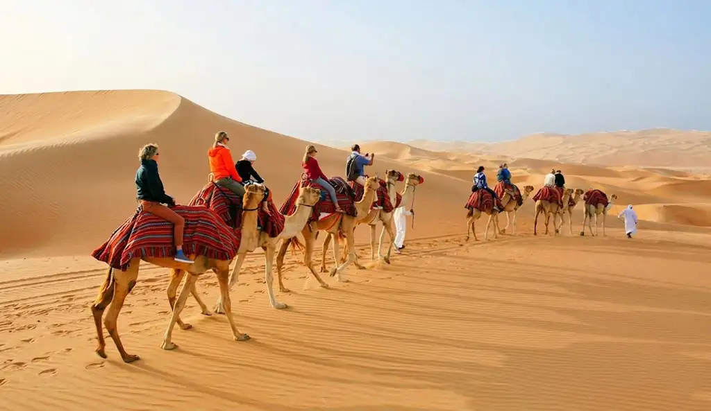 A large group caravan of travelers camel trekking across rolling desert sand dunes during an Abu Dhabi morning safari adventure.