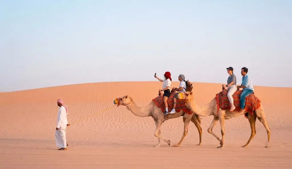 Tourists enjoying a scenic camel ride in the golden sand dunes of Abu Dhabi desert, led by a local guide in traditional Emirati attire during a sunset camel trekking experience.