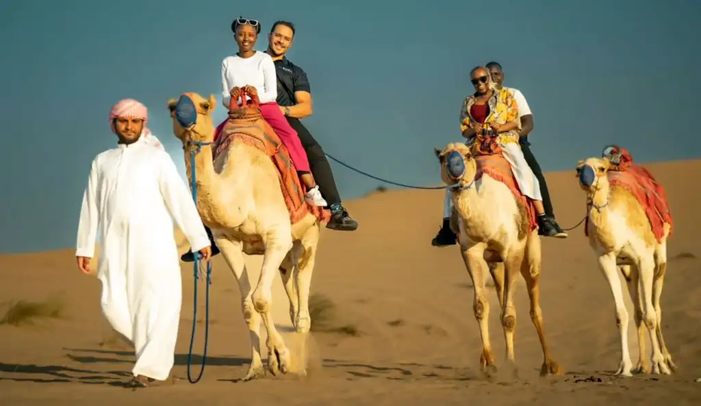 A group of smiling tourists riding camels through the golden sand dunes of Abu Dhabi Desert on a bright sunny morning. A local guide in traditional white kandura and ghutra leads the caravan, with riders enjoying the scenic desert landscape under a clear blue sky.