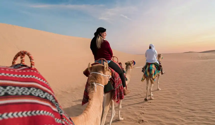 A woman and a man in a white thobe ride camels through endless Abu Dhabi golden sand dunes.