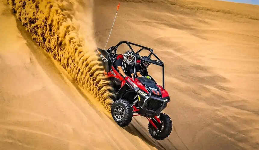 Red Polaris RZR dune buggy powering up a steep sand dune with massive sand spray during an exciting dune buggy tour in Abu Dhabi desert.