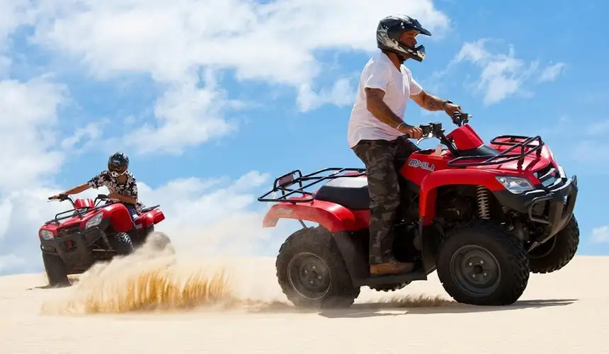 Exciting quad bike tour in Abu Dhabi desert with two riders on powerful red ATVs racing across golden sand dunes, kicking up sand under a bright blue sky. Perfect desert adventure experience in Abu Dhabi.