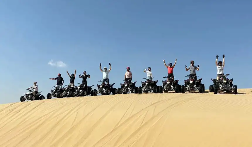 Happy group of adventurers celebrating on quad bikes at the top of a massive sand dune during a quad bike tour in Abu Dhabi desert. Unforgettable desert safari experience with stunning views.