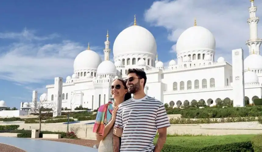 Smiling couple posing in front of the majestic white domes and minarets of the Sheikh Zayed Grand Mosque in Abu Dhabi on a sunny day.
