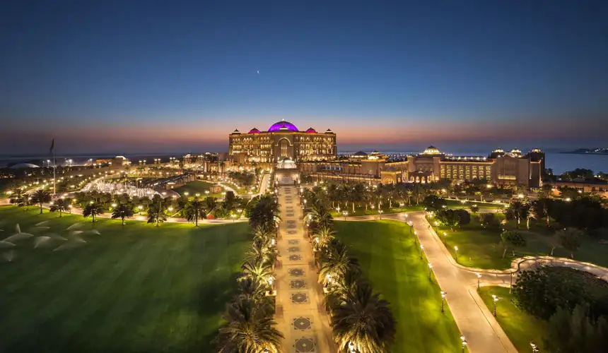 Stunning nighttime view of the luxurious Emirates Palace hotel in Abu Dhabi, illuminated with purple domes, fountains, and lush gardens along the waterfront.