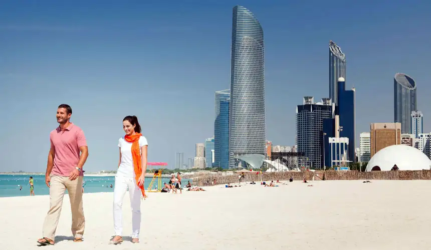 A happy couple strolling along a sandy beach in Abu Dhabi with the iconic twisted skyscraper and modern skyline in the background under a clear blue sky.