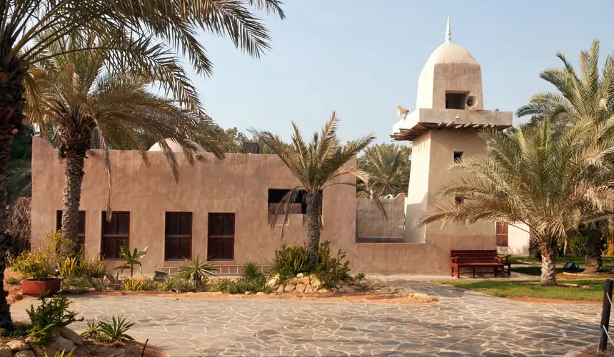 Traditional mud-brick Arabian house with a small minaret and dome surrounded by date palm trees in a desert setting under clear sky.
